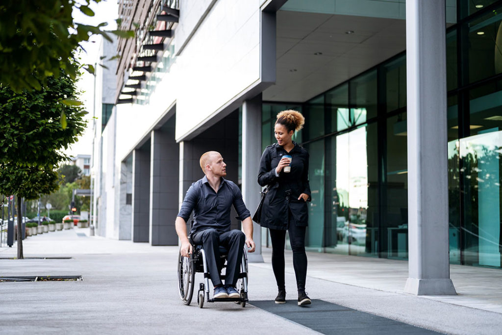 A white man in a wheelchair and an African American woman walk past buildings on their way to work.