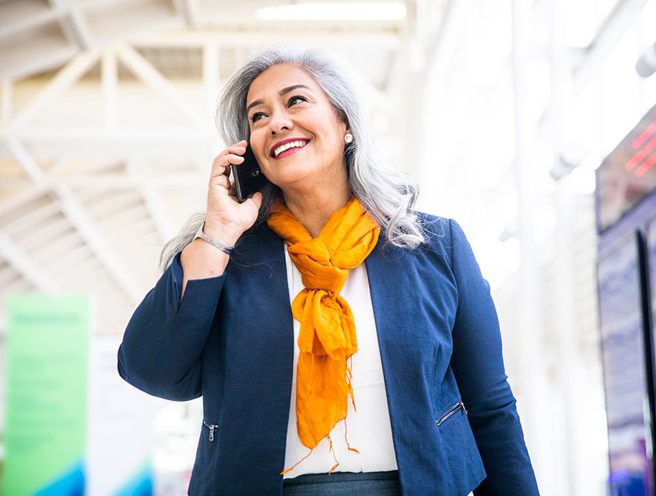 Mature woman in a blue jacket and orange scarf happily talks on her cellphone.
